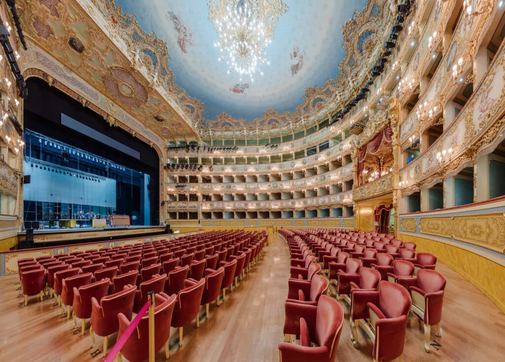 A view from the side of Teatro La Fenice, showing the theatre's red seats, ornate balconies, beautiful blue ceiling and chandelier. The stage is to the right.