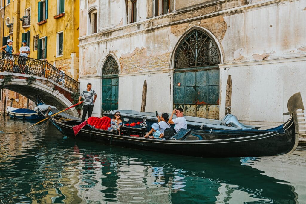 A group of four tourists enjoy a gondola ride in Venice, steered by a man in a striped t-shirt. Taking a gondola ride is one of the best activities to do in Venice in January.