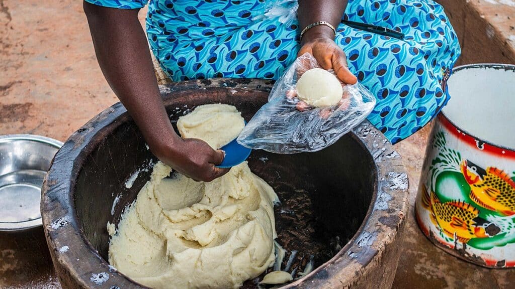 A person in a blue dress making pounded yam from a bucket. Pounded yam is one of the national foods of Nigeria.