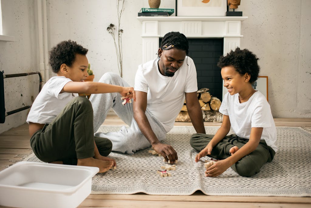 Full body of cheerful African twin brothers in similar casual clothes smiling while sitting on floor and playing with toys together with happy young father