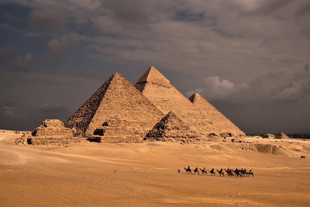 In the distance, the iconic pyramids of Giza rise majestically against the sky, framed by the golden hues of the desert. A caravan of camels leisurely makes its way towards the ancient wonders.