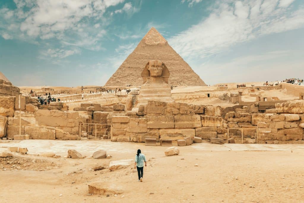 A man walks toward the Sphinx in Giza, with the majestic backdrop of The Pyramids of Giza towering in the distance, embodying the timeless wonder and grandeur of ancient Egypt.